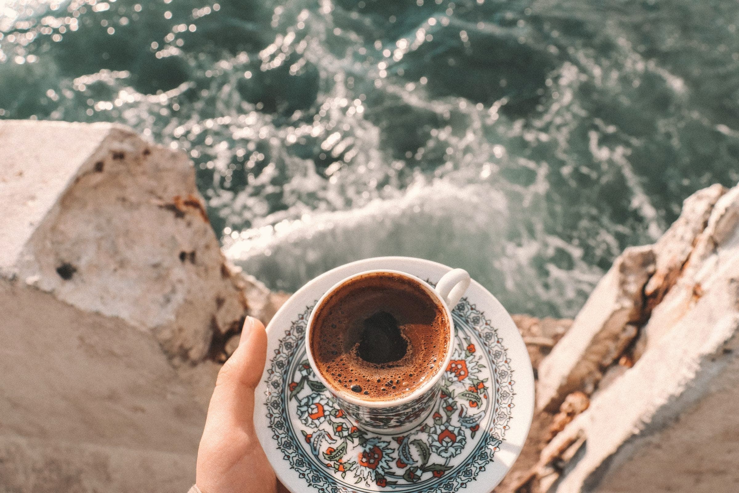 Turkish coffee in a patterned cup held above breaking sea waves