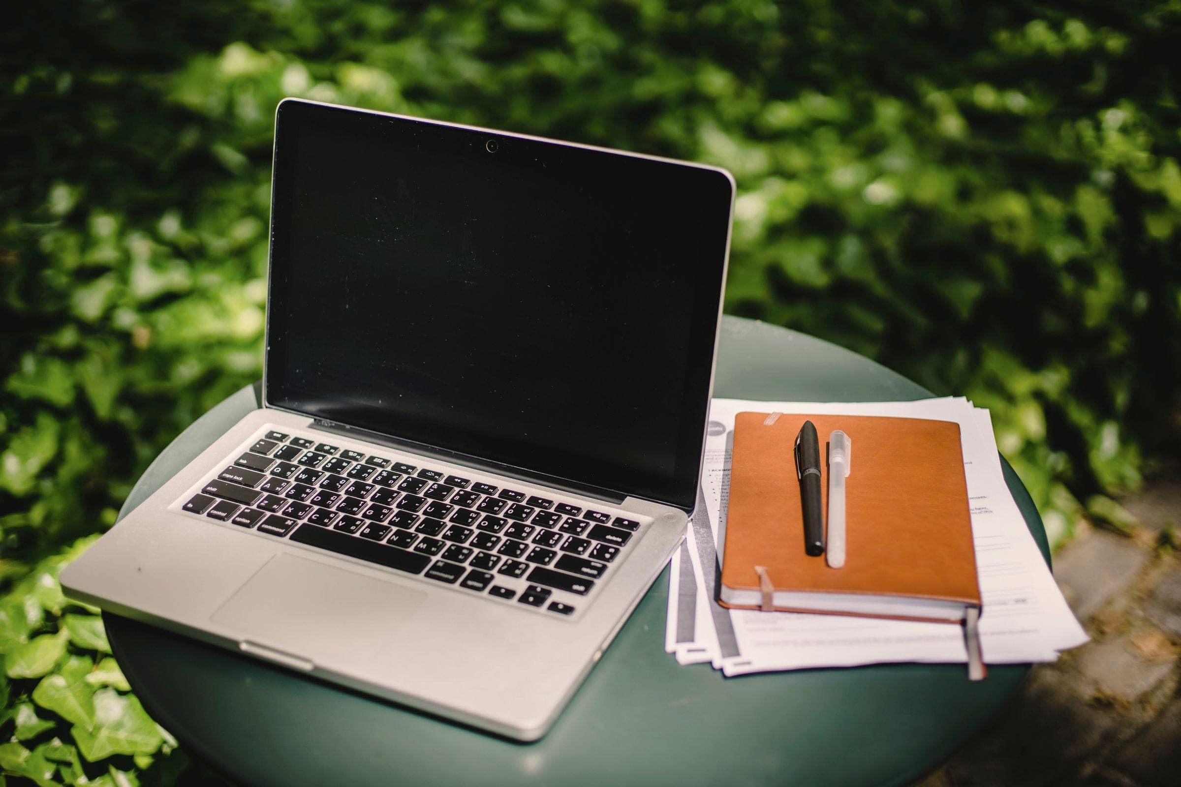 An open laptop with a notebook and pen on a round table in a green garden