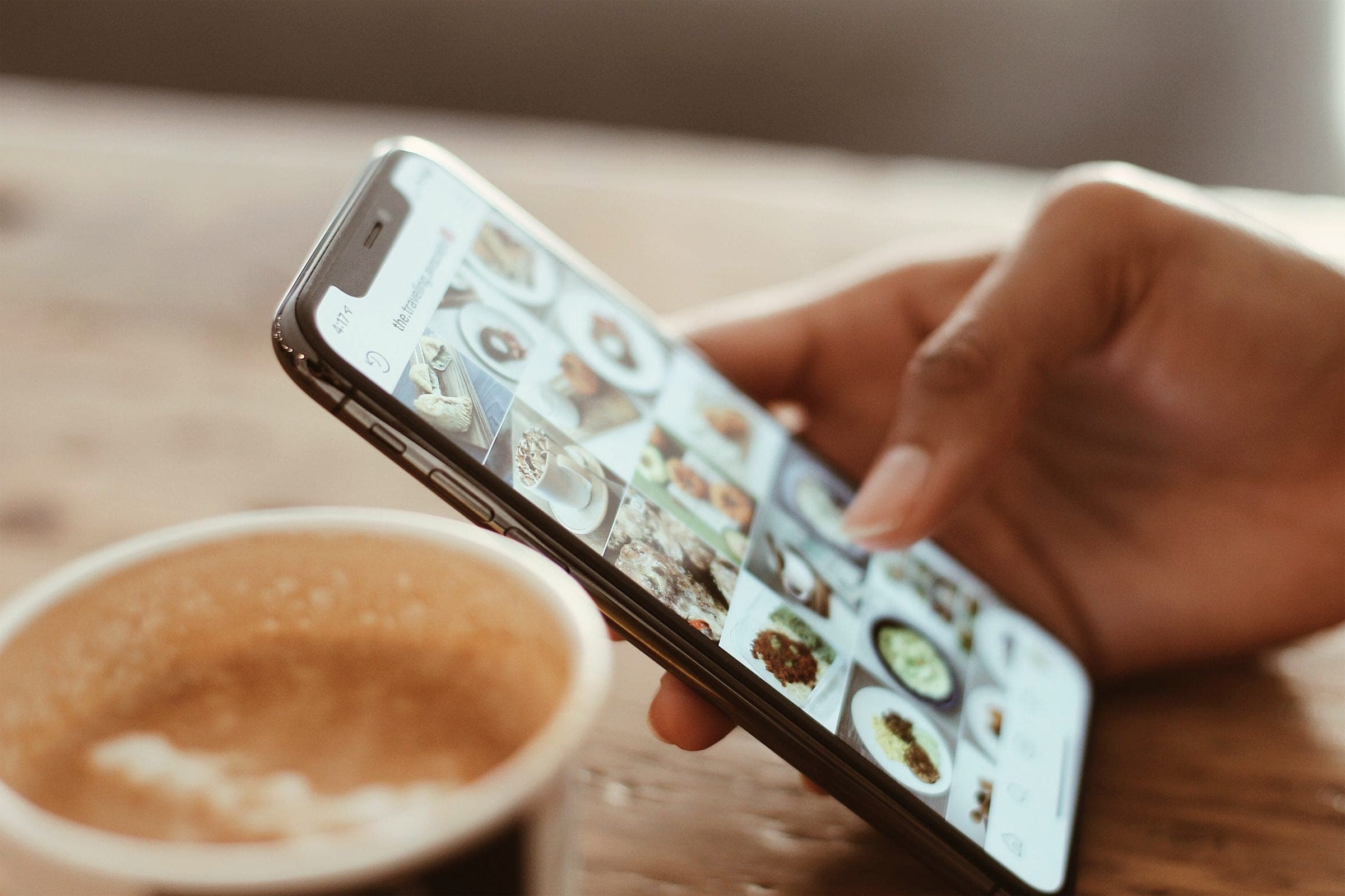 A hand scrolling through a photo feed on a phone next to a coffee cup