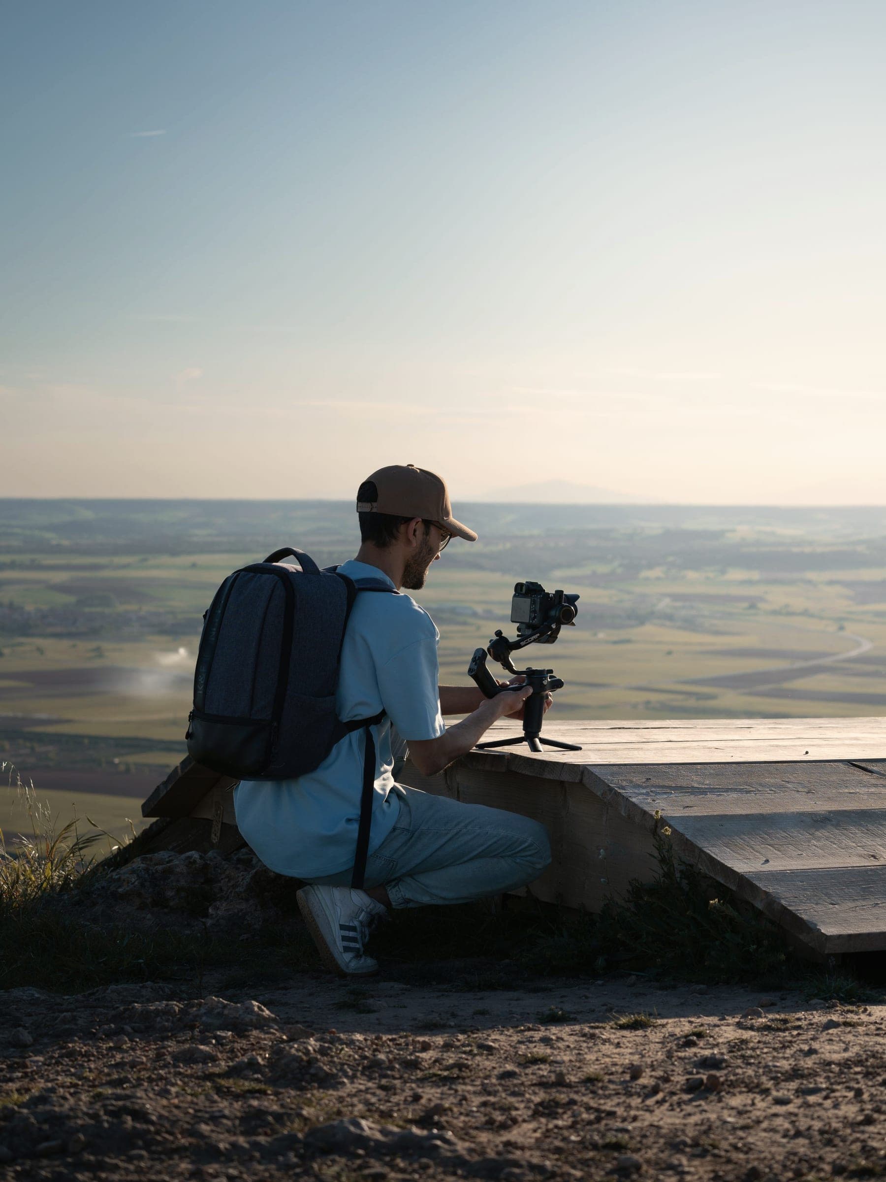 A person with a backpack kneels on a hilltop, filming a distant plain with a gimbal