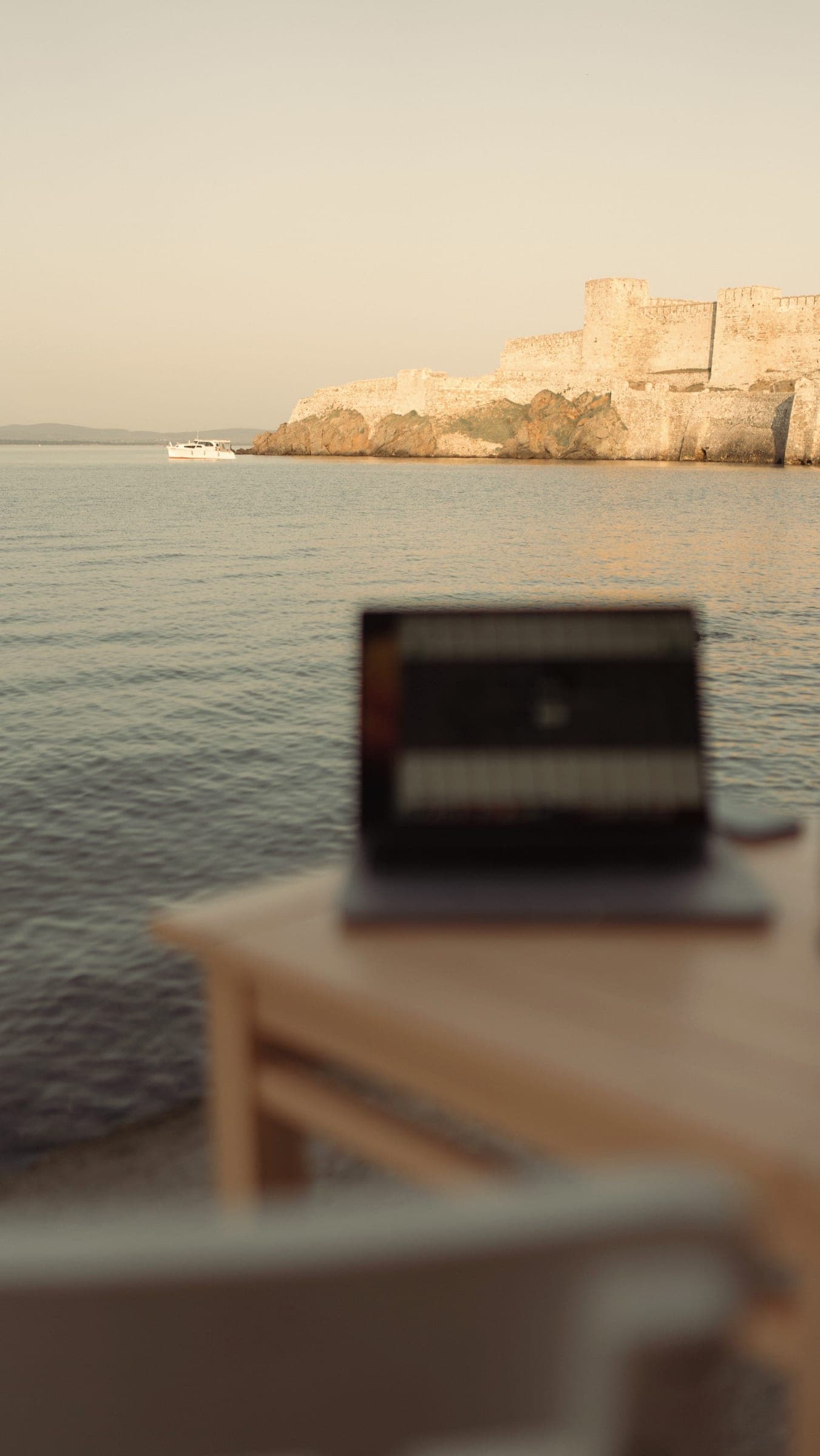 A laptop on a table by the shore with a rocky headland behind (Bozcaada)