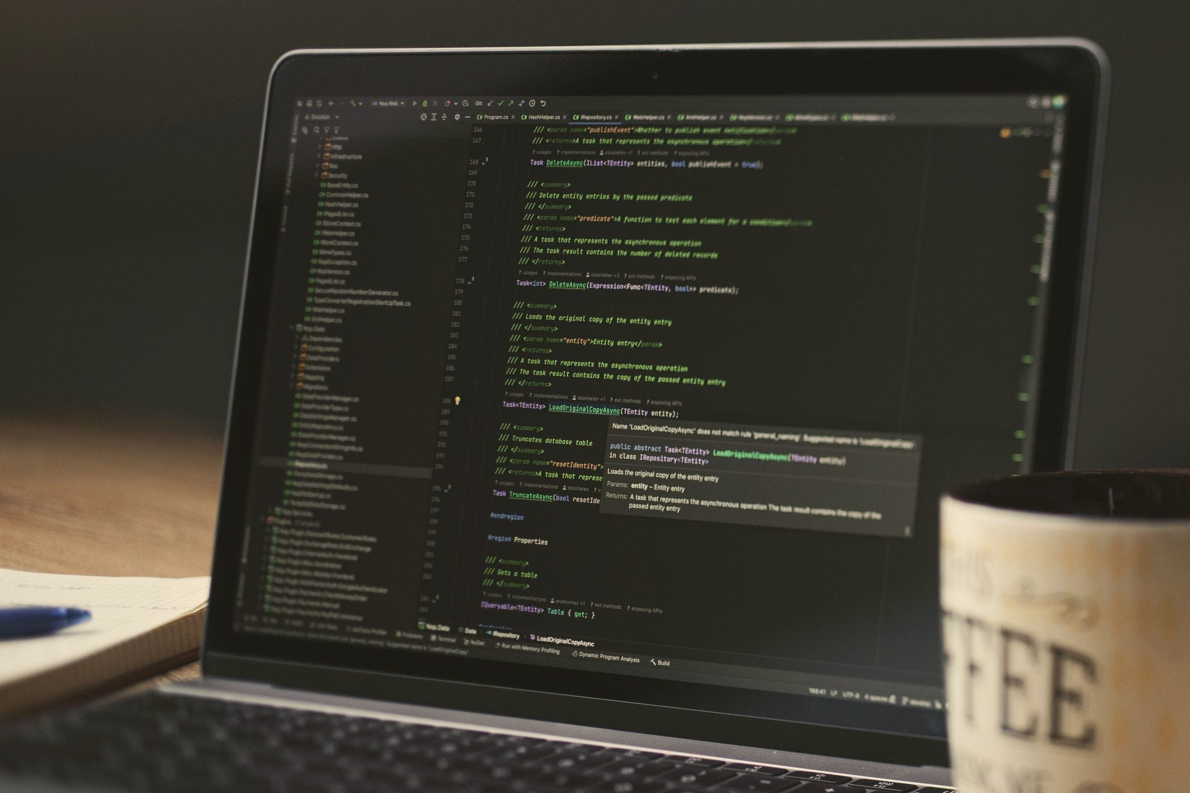 A laptop showing code in a dark editor theme, with a mug beside it