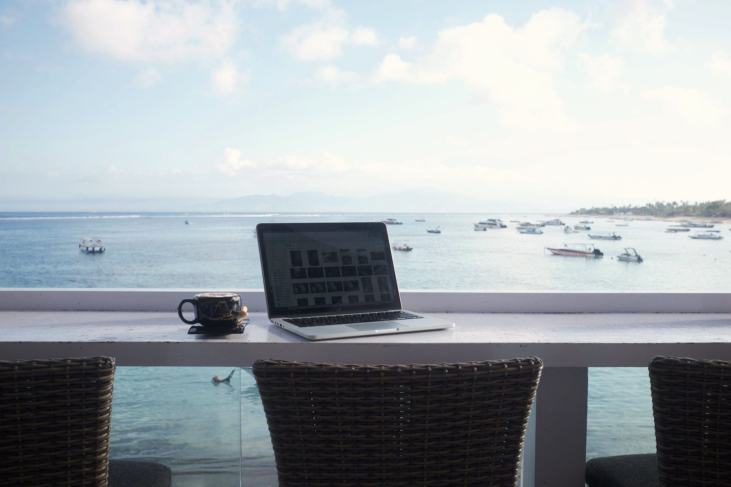 A laptop and coffee cup on a bar counter by a window overlooking the sea and boats